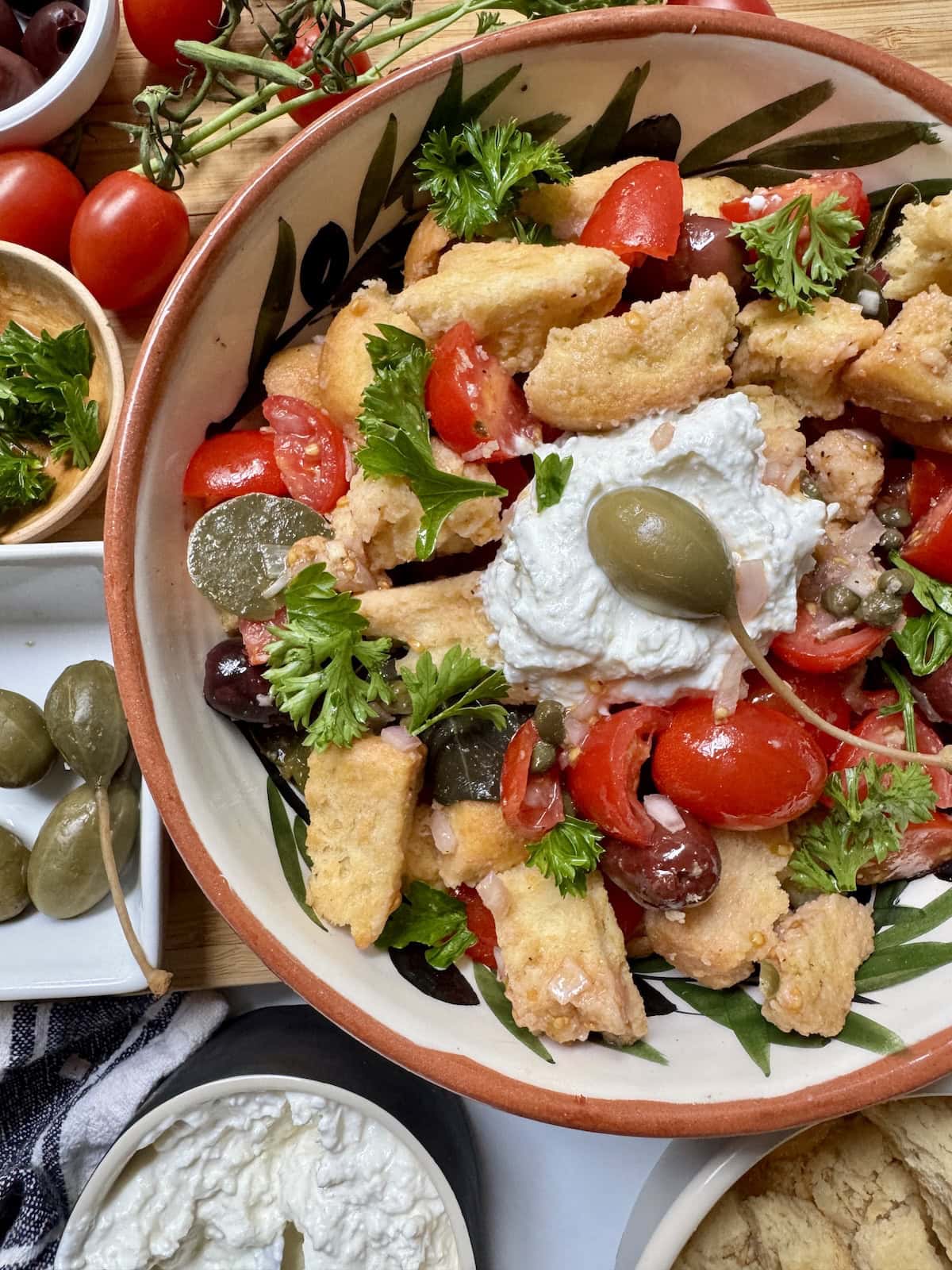 Salad with tomatoes, parsley, croutons, capers, and whipped feta cheese in a bowl with small bowls of ingredients to the side.