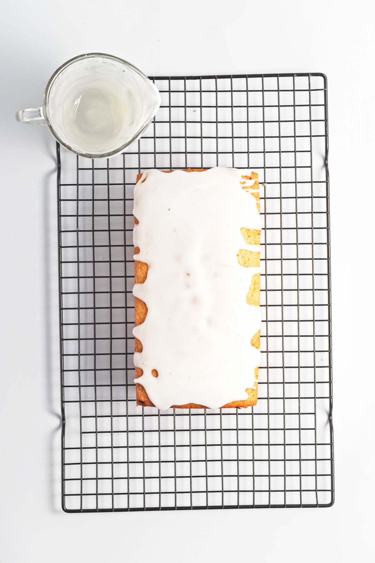Loaf cake on wire rack with empty measuring cup of icing.