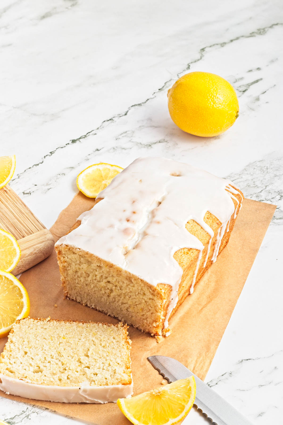 Lemon loaf cake on parchment paper with lemon pieces scattered on wood cutting board and whole lemon in background.
