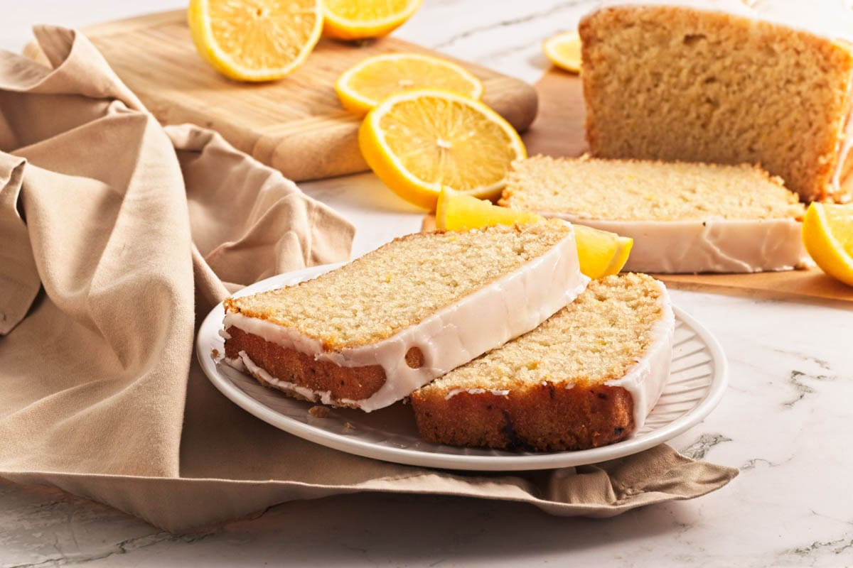 Lemon loaf cake slice on plate with lemon pieces scattered on wood cutting board.
