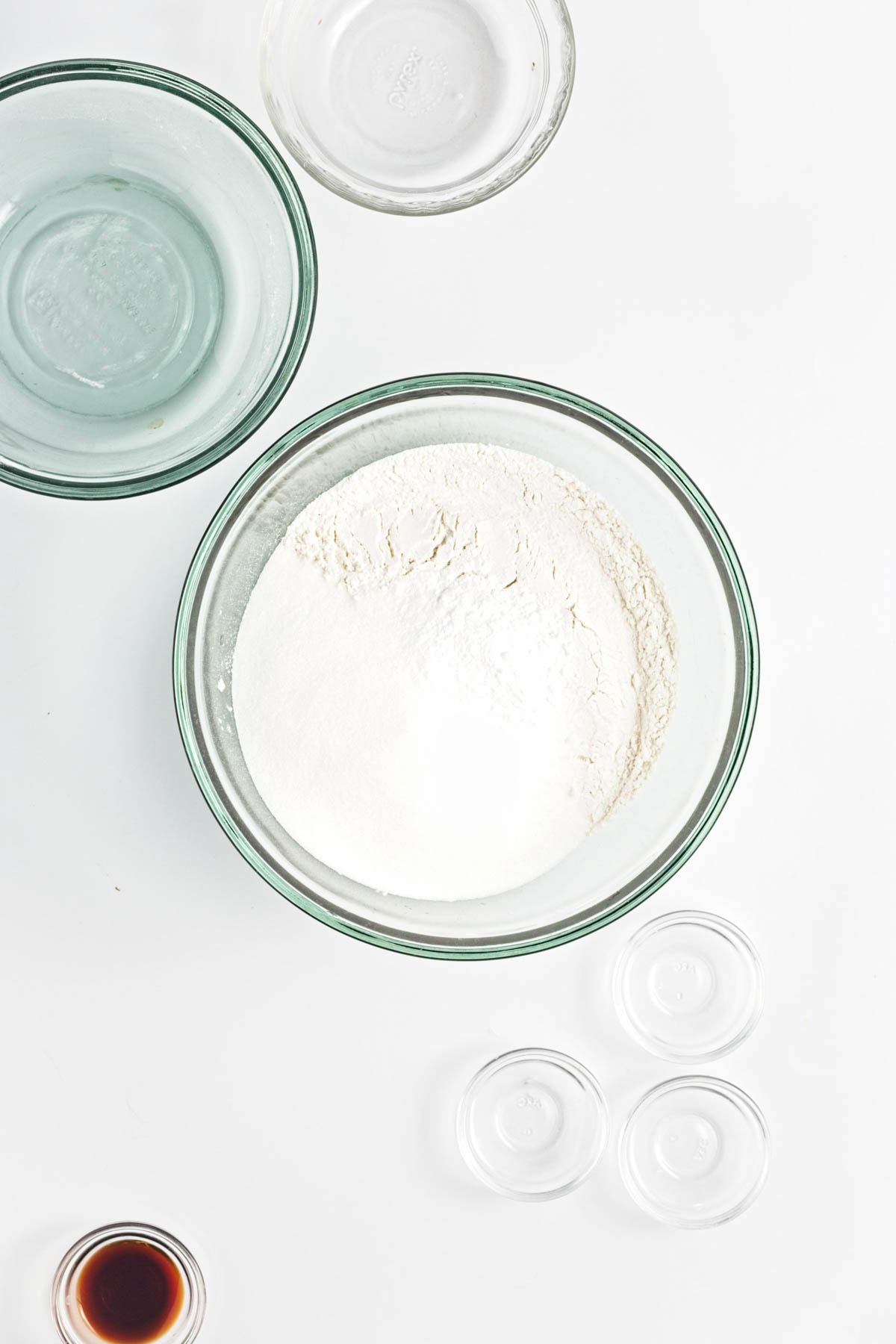 Glass bowl of flour and other dry ingredients on white counter with empty bowls and small bowl of vanilla.