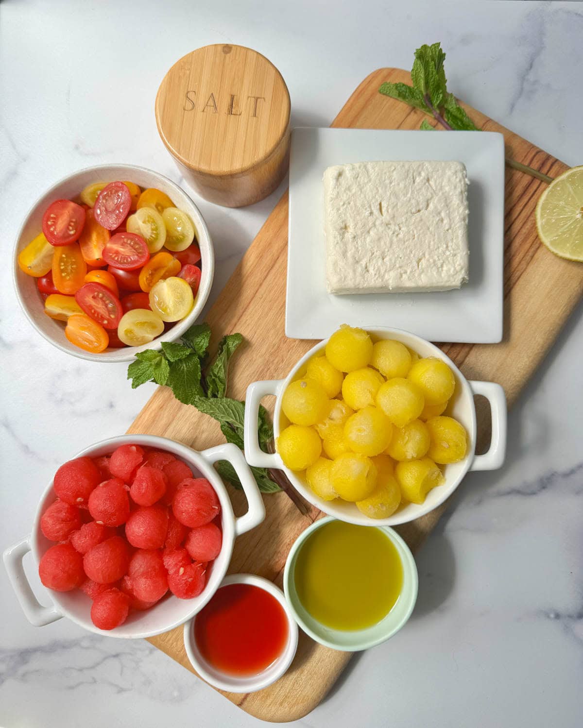 Bowls of tomatoes, watermelon, olive oil, vinegar, and a block of feta cheese with sprigs of mint and a lime half.