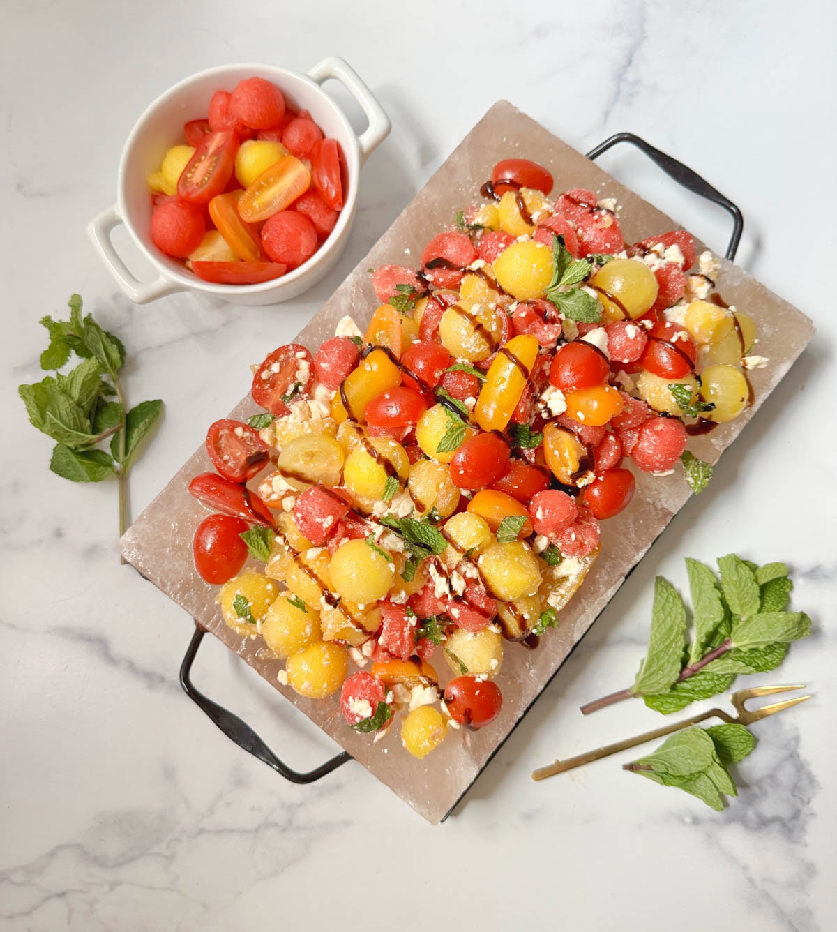 A platter of watermelon salad with a bowl of tomatoes and watermelon balls with fresh mint on the side.