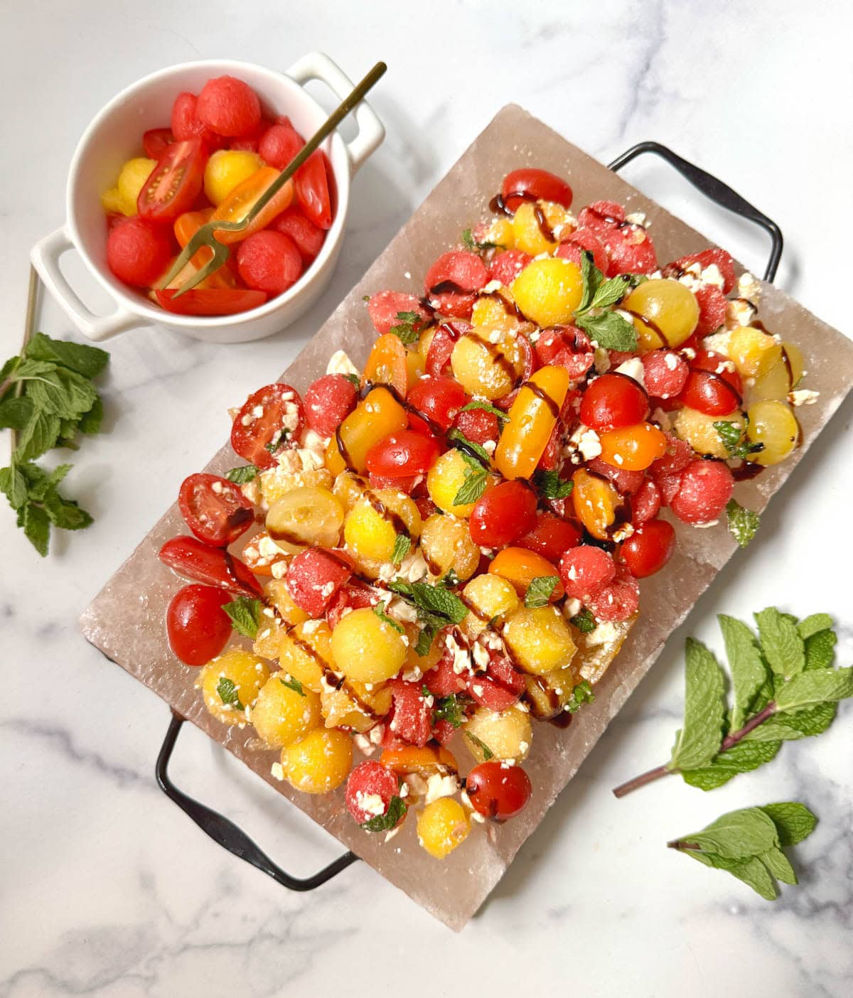 A platter of watermelon salad with a bowl of tomatoes and watermelon balls with fresh mint on the side.