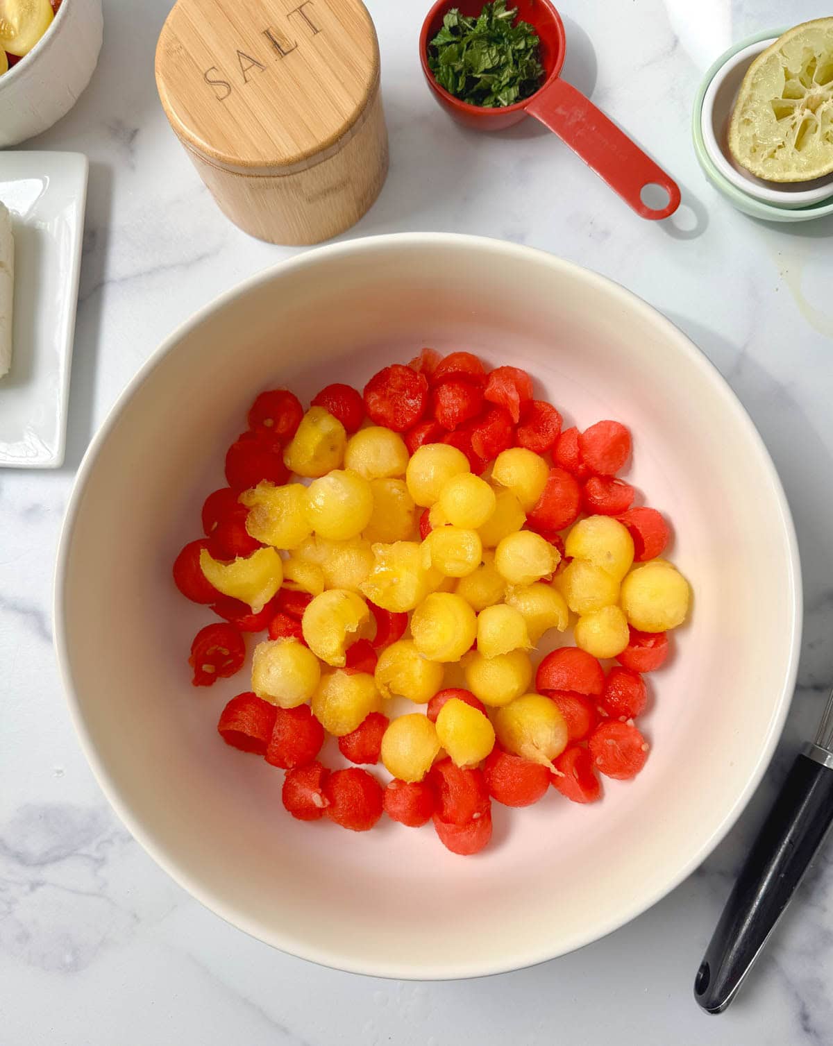 White bowl with red and yellow watermelon balls, a salt container, cup of mint, and lime above.
