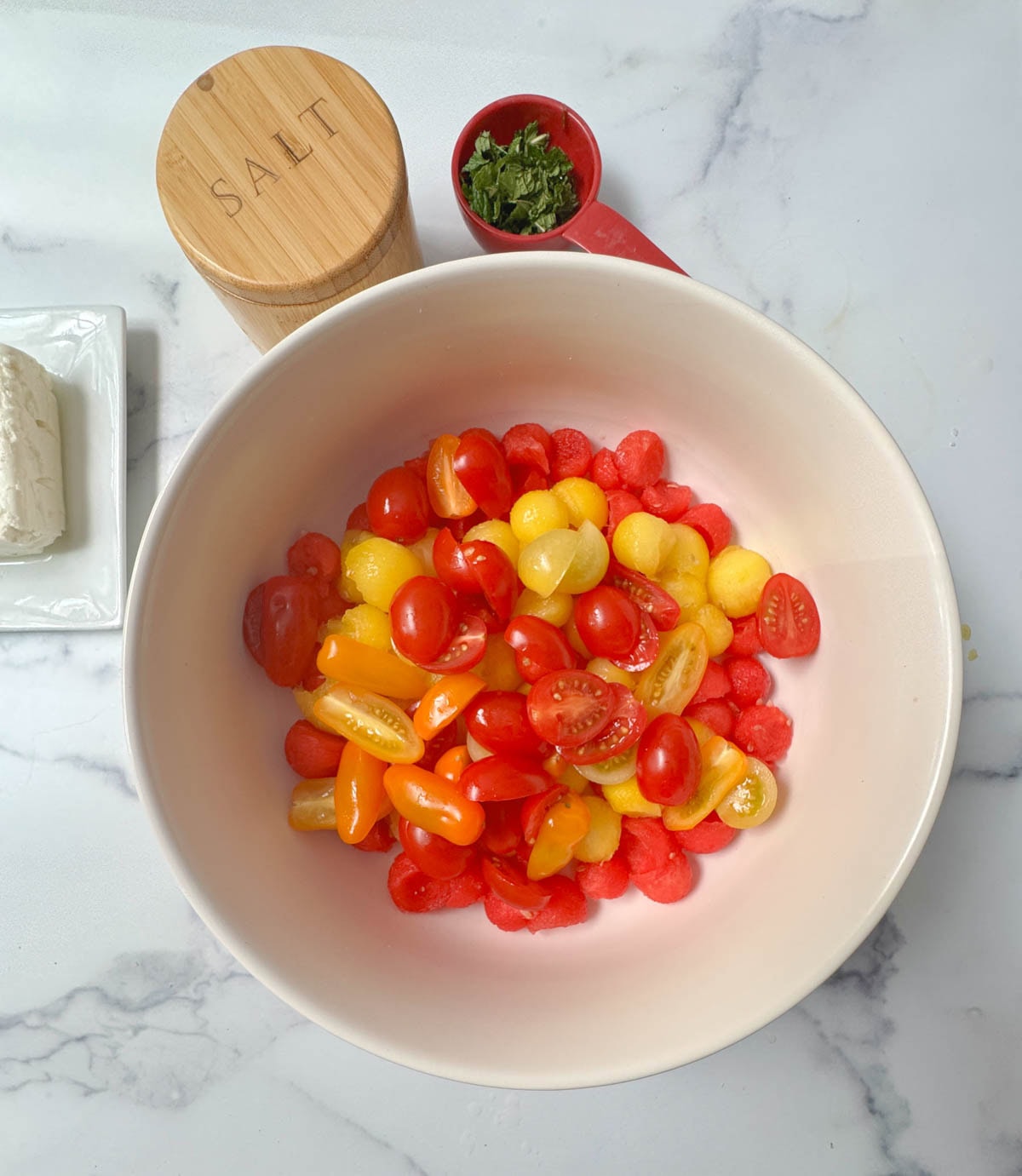 White bowl of red and yellow tomato halves with cup of mint, salt container, and feta cheese to the side.
