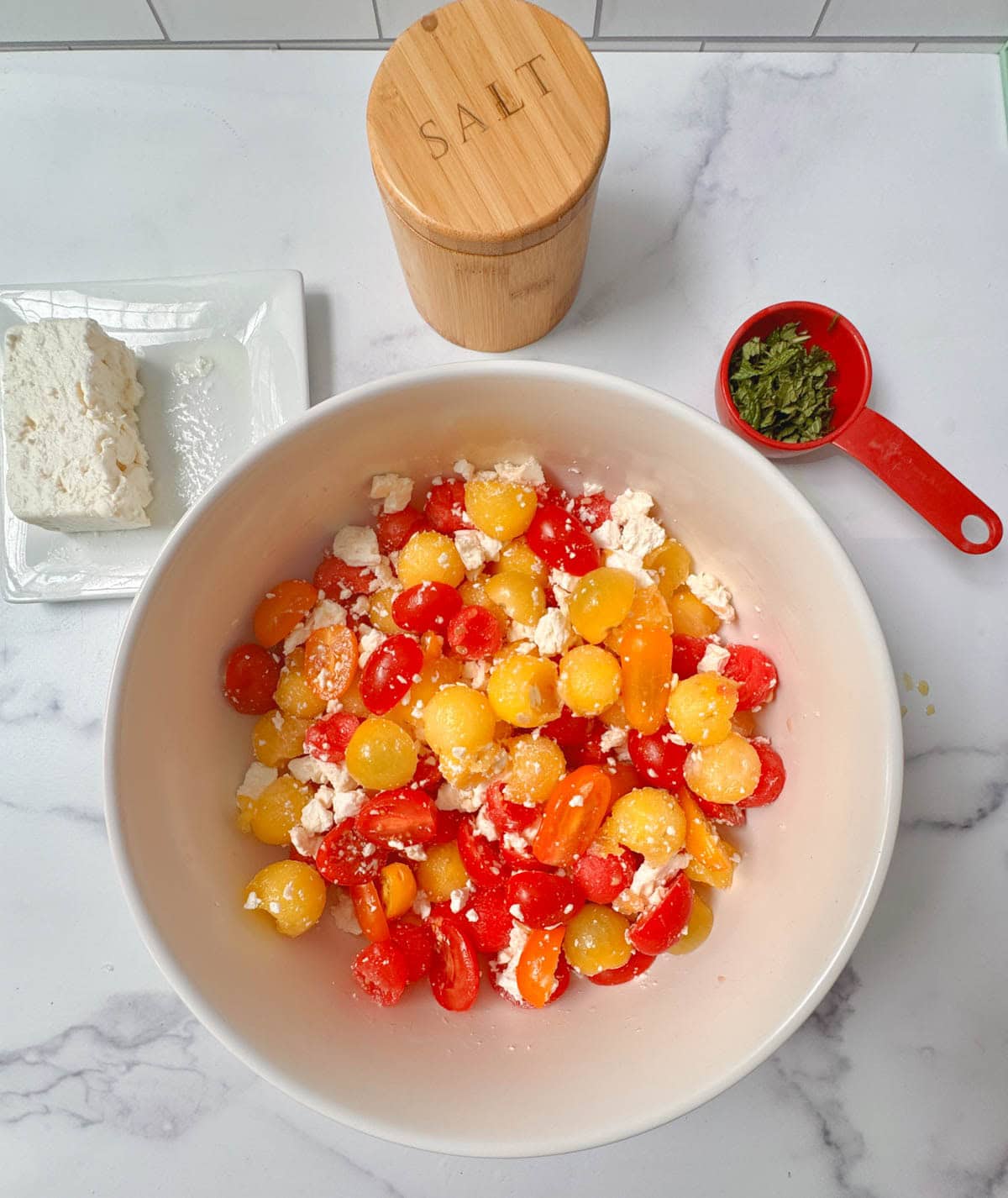 White bowl of red and yellow tomato halves, red and yellow watermelon balls, and feta cheese, with cup of mint, and salt container to the side.
