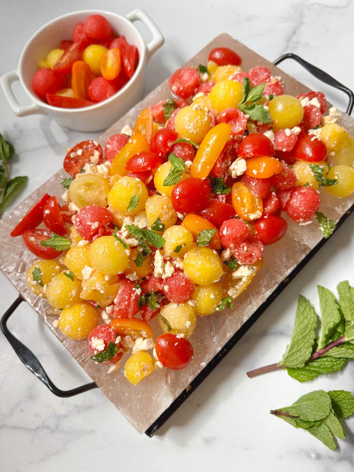 A platter of watermelon salad with a bowl of tomatoes and watermelon balls with fresh mint on the side.