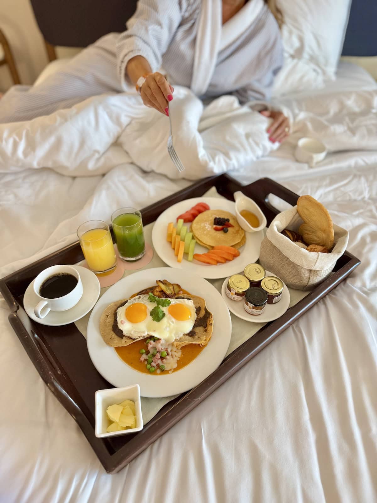 Tray of breakfast food on a bed with woman in background with a fork.