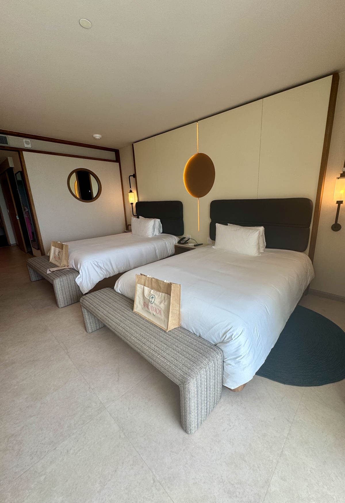 Two queen hotel beds with bench in front and bags on bench.