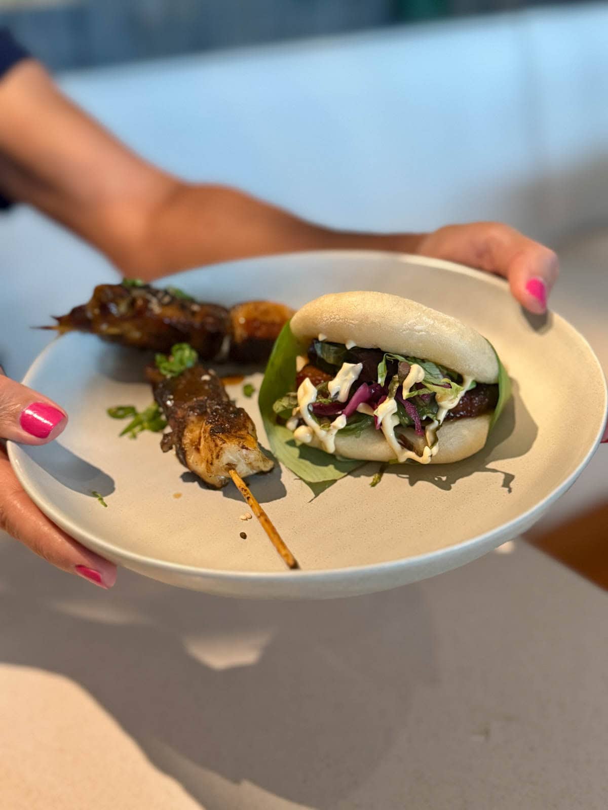 Hands holding white bowl with bao bun and chicken skewers.
