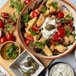 Bowl with salad of tomatoes, capers, shallots, olives, parsley, and dressing topped with feta cheese mixture and caperberry.