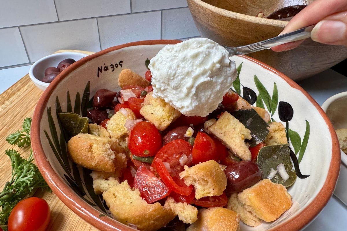 Placing spoonful of creamy feta on a salad of tomatoes, croutons, olives, shallots, capers, and caper leaves.