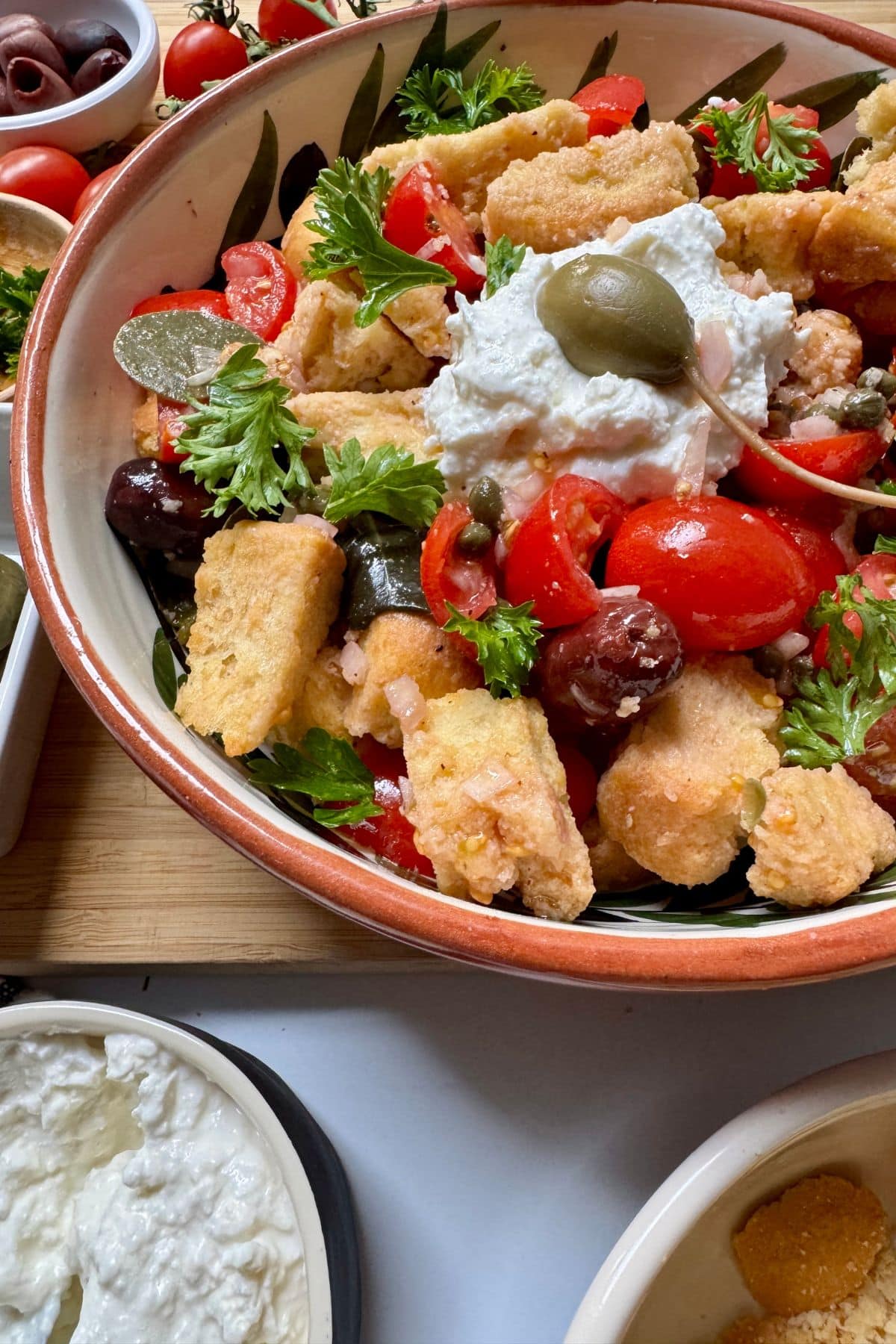 Salad with tomatoes, parsley, croutons, capers, and whipped feta cheese in a bowl with small bowls of ingredients to the side.