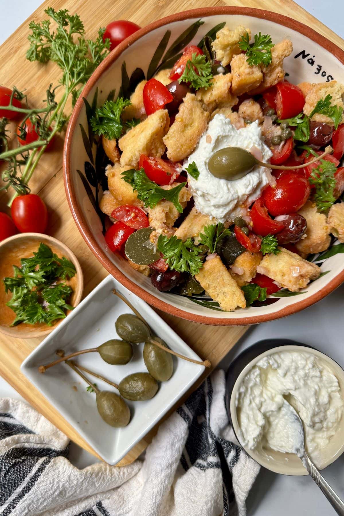 Salad with tomatoes, parsley, croutons, capers, and whipped feta cheese in a bowl with small bowls of ingredients to the side.