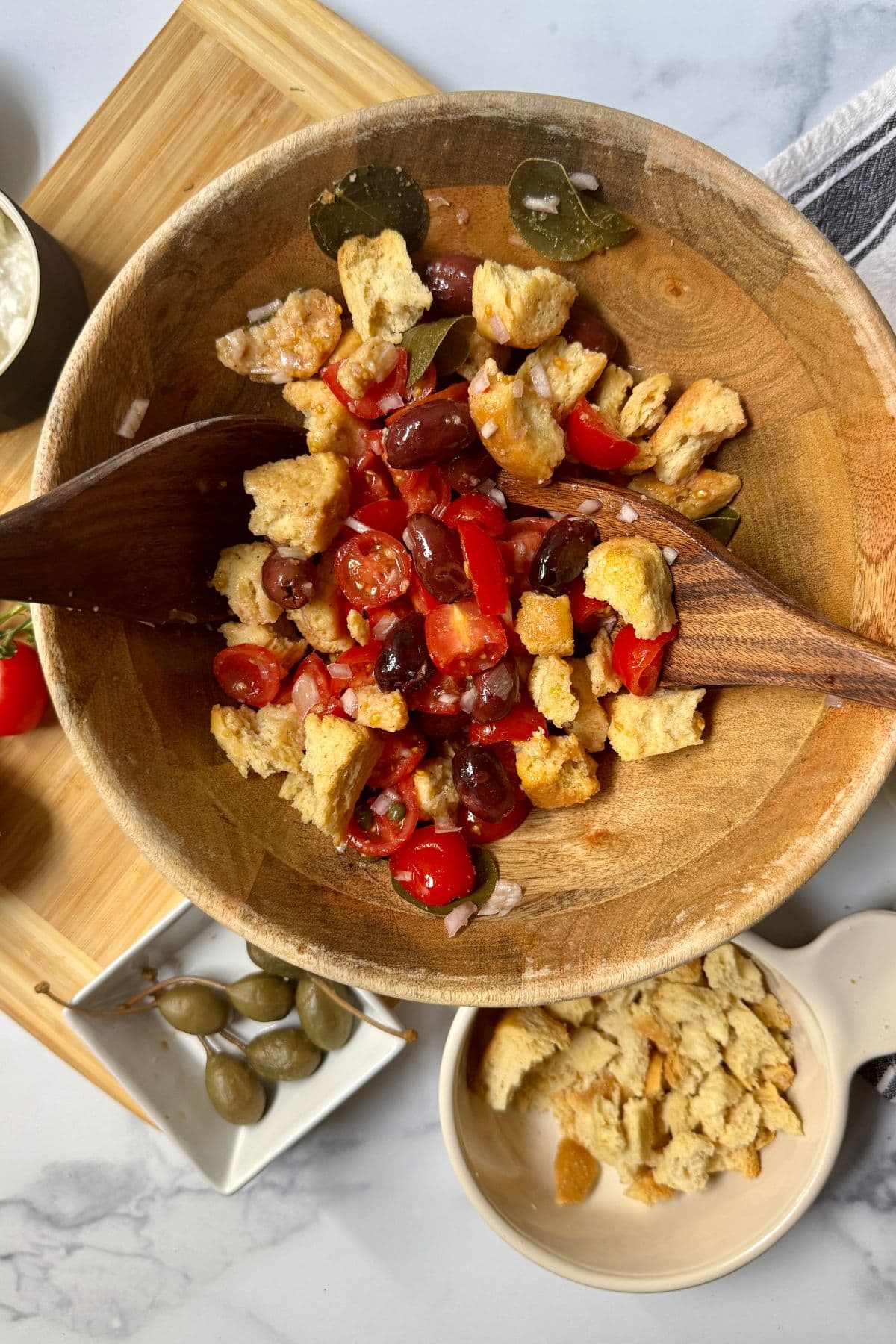 Wooden bowl with tomatoes cut in half, chopped shallots, kalamata olives, capers, caper leaves, and barley rusks,