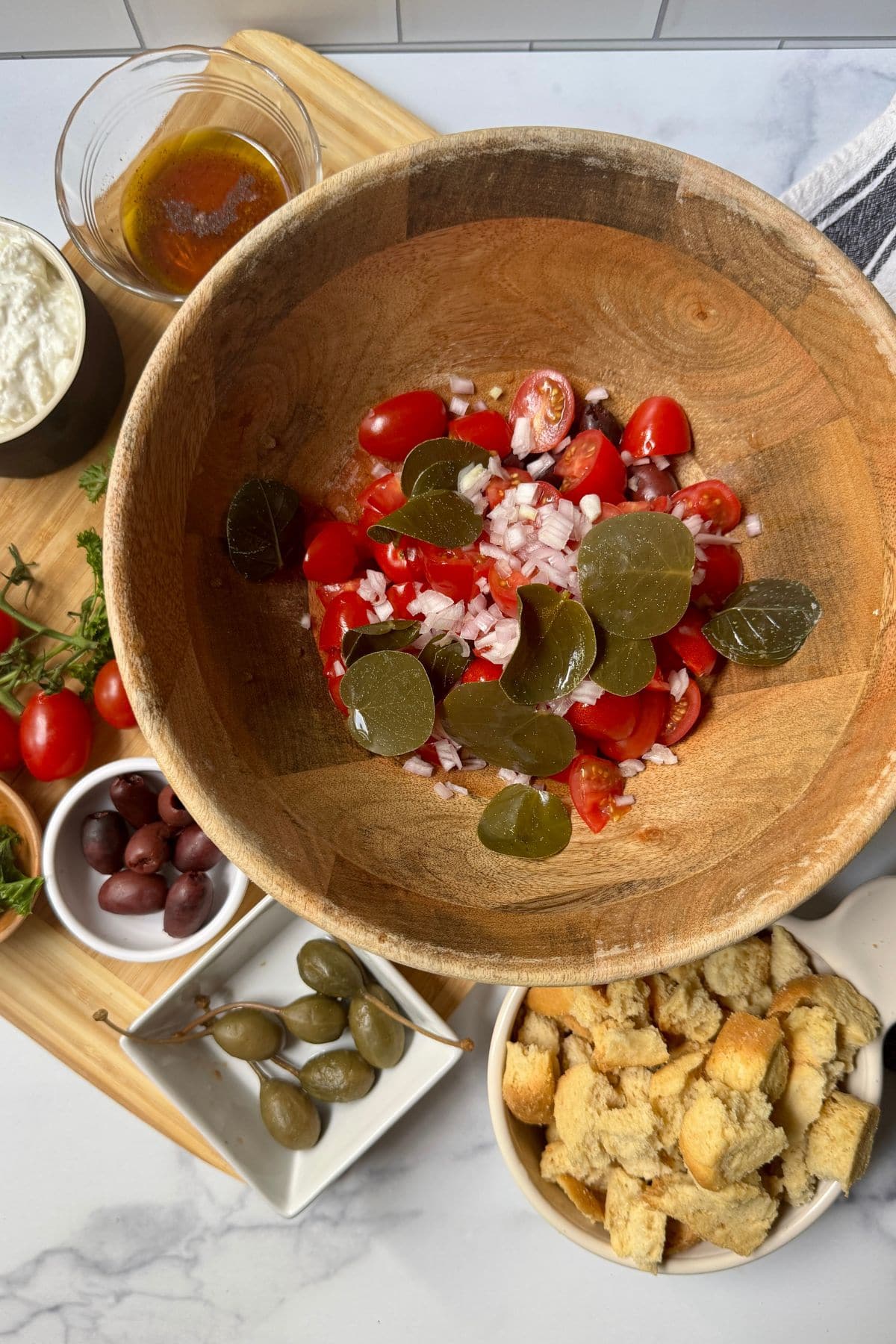Wooden bowl with tomatoes cut in half, chopped shallots, kalamata olives and caper leaves.