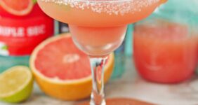 Cocktail set on a wood board made with grapefruit juice in a margarita glass rimmed with salt with lime slices in drink and grapefruit slice on side of rim. Bottle of liqueur in background.