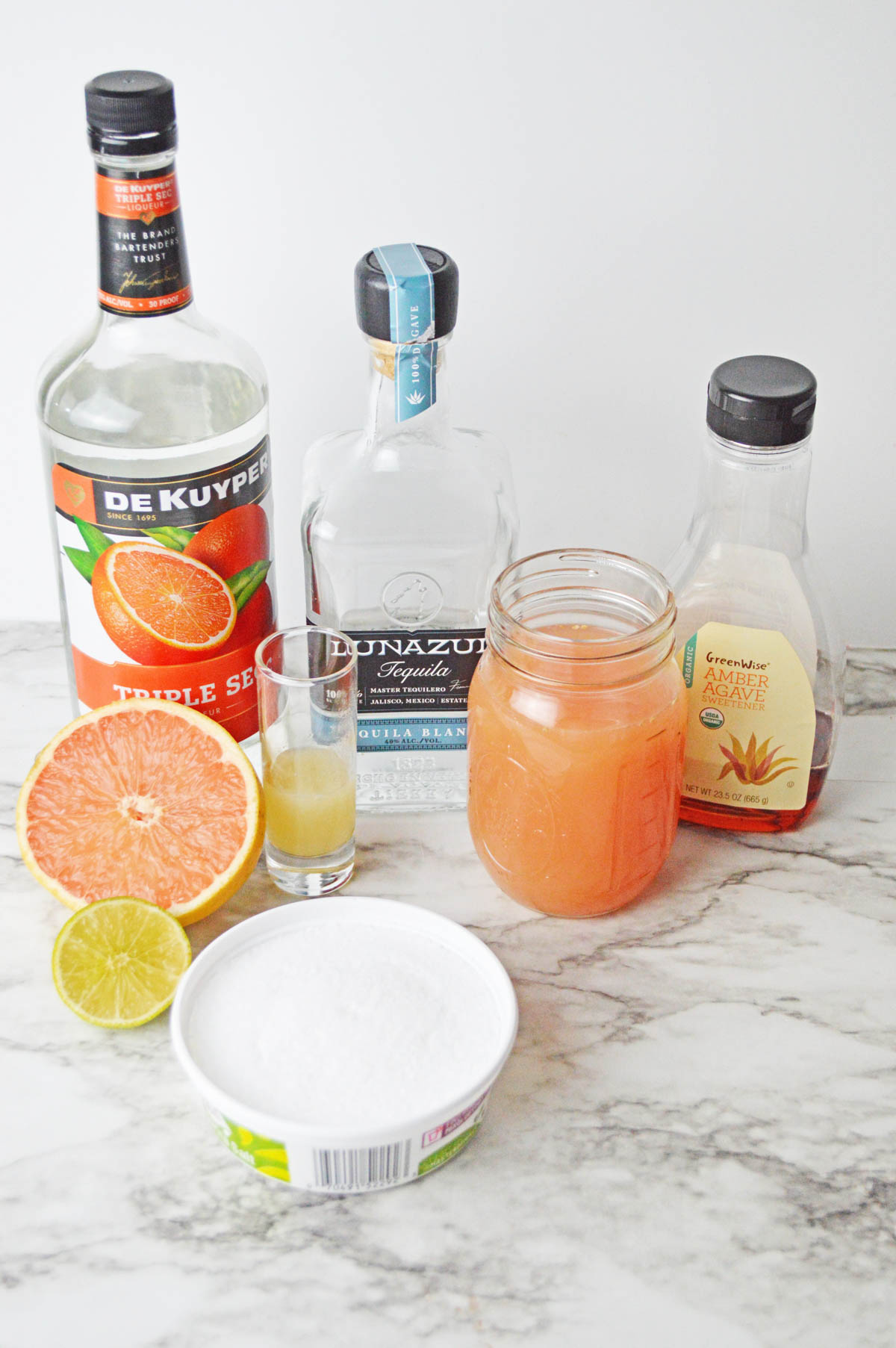 Bottle of orange liqueur, tequila, and agave with a mason jar of grapefruit juice, shot glass of lime juice, container of salt, and fresh half grapefruit and half lime on marble counter.
