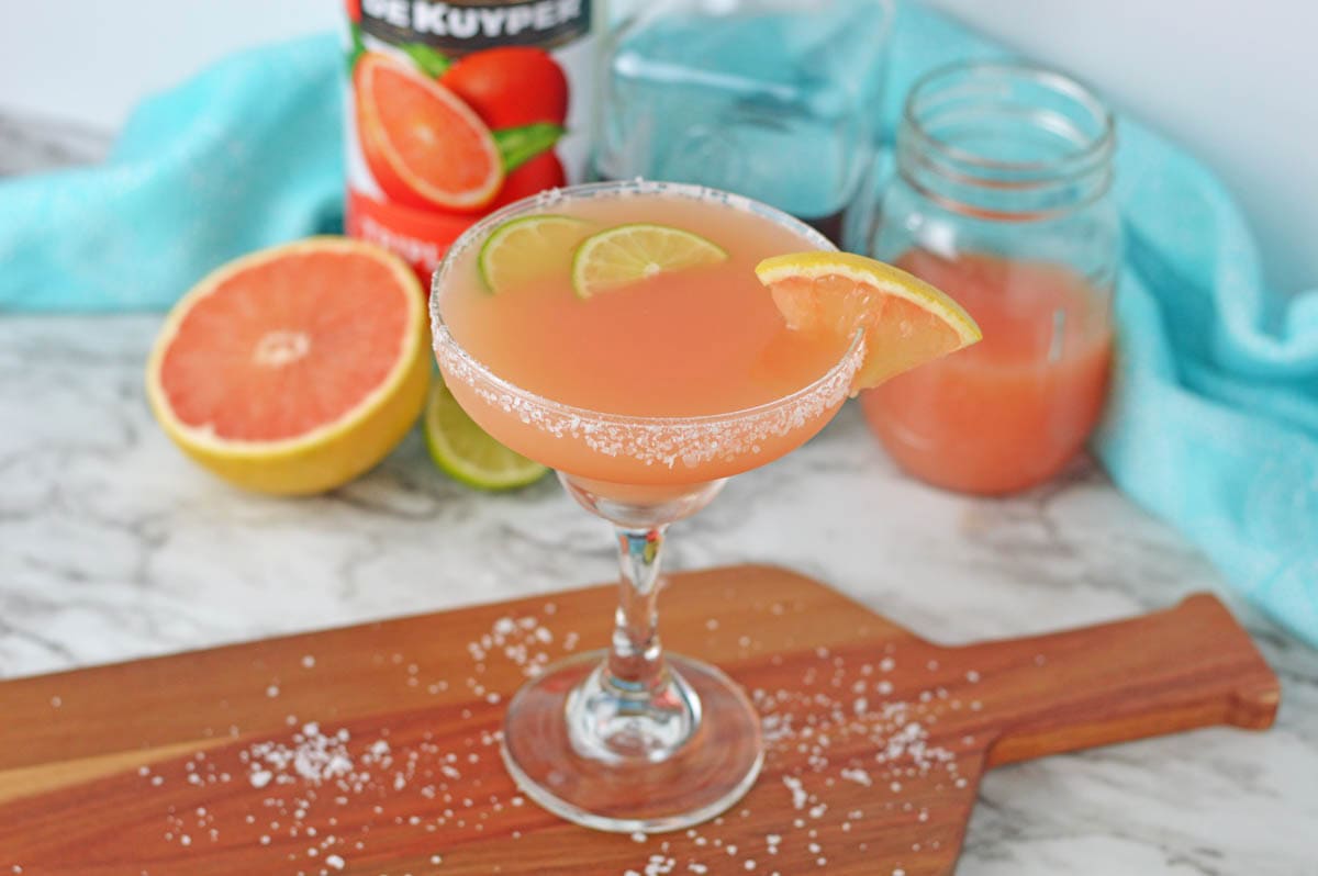 Cocktail set on a wood board made with grapefruit juice in a margarita glass rimmed with salt with lime slices in drink and grapefruit slice on side of rim. Bottle of liqueur in background.