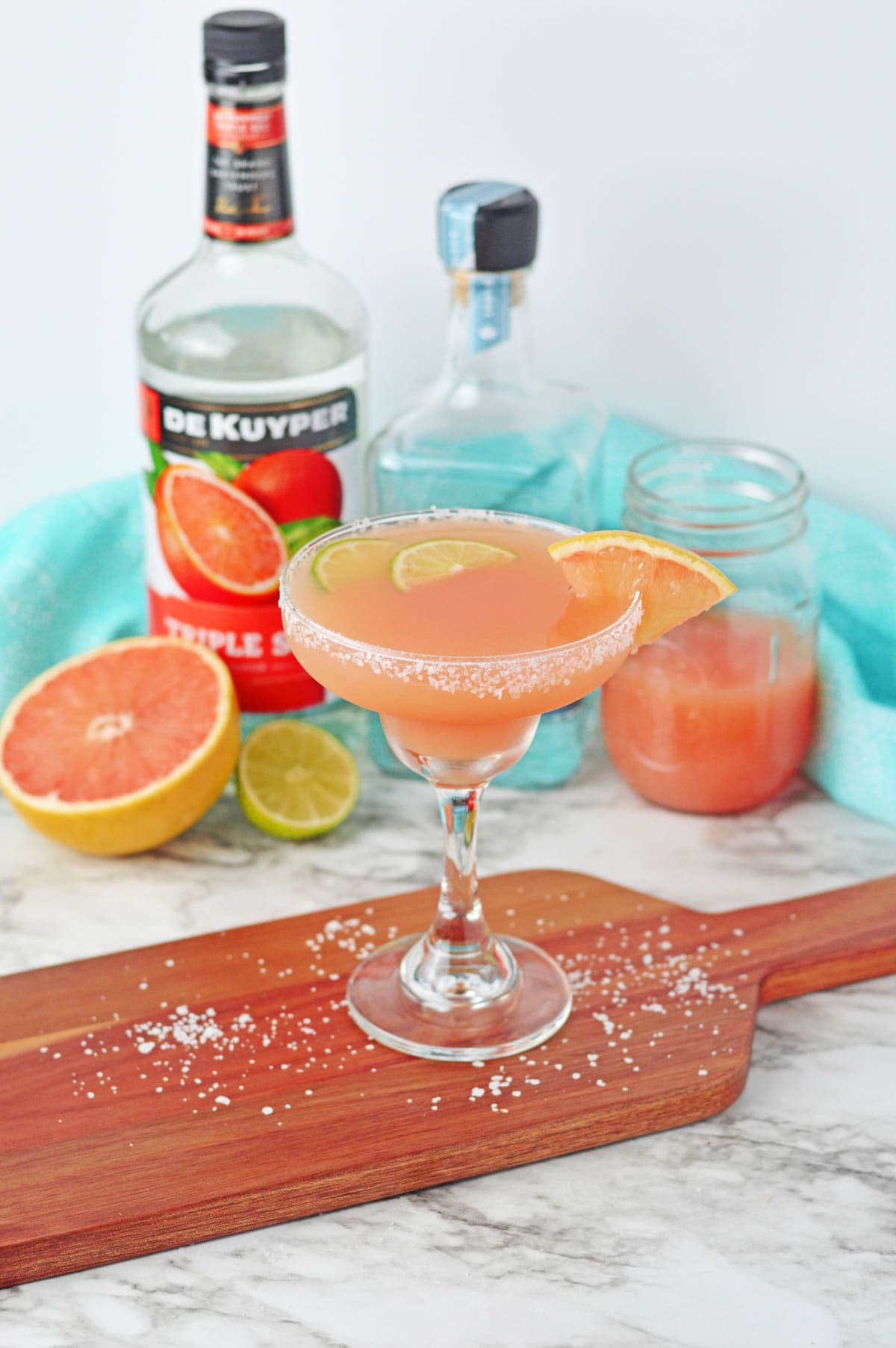 Cocktail set on a wood board made with grapefruit juice in a margarita glass rimmed with salt with lime slices in drink and grapefruit slice on side of rim. Bottle of liqueur in background.