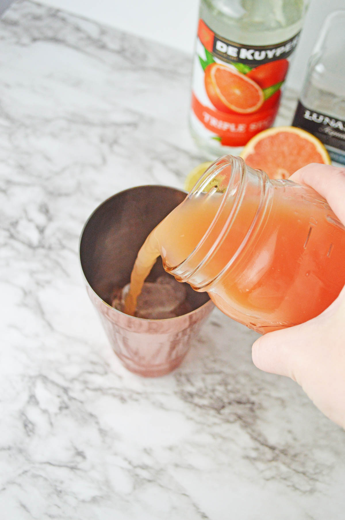 Pouring grapefruit juice into a cocktail shaker with grapefruit and bottle of orange liqueur in background.