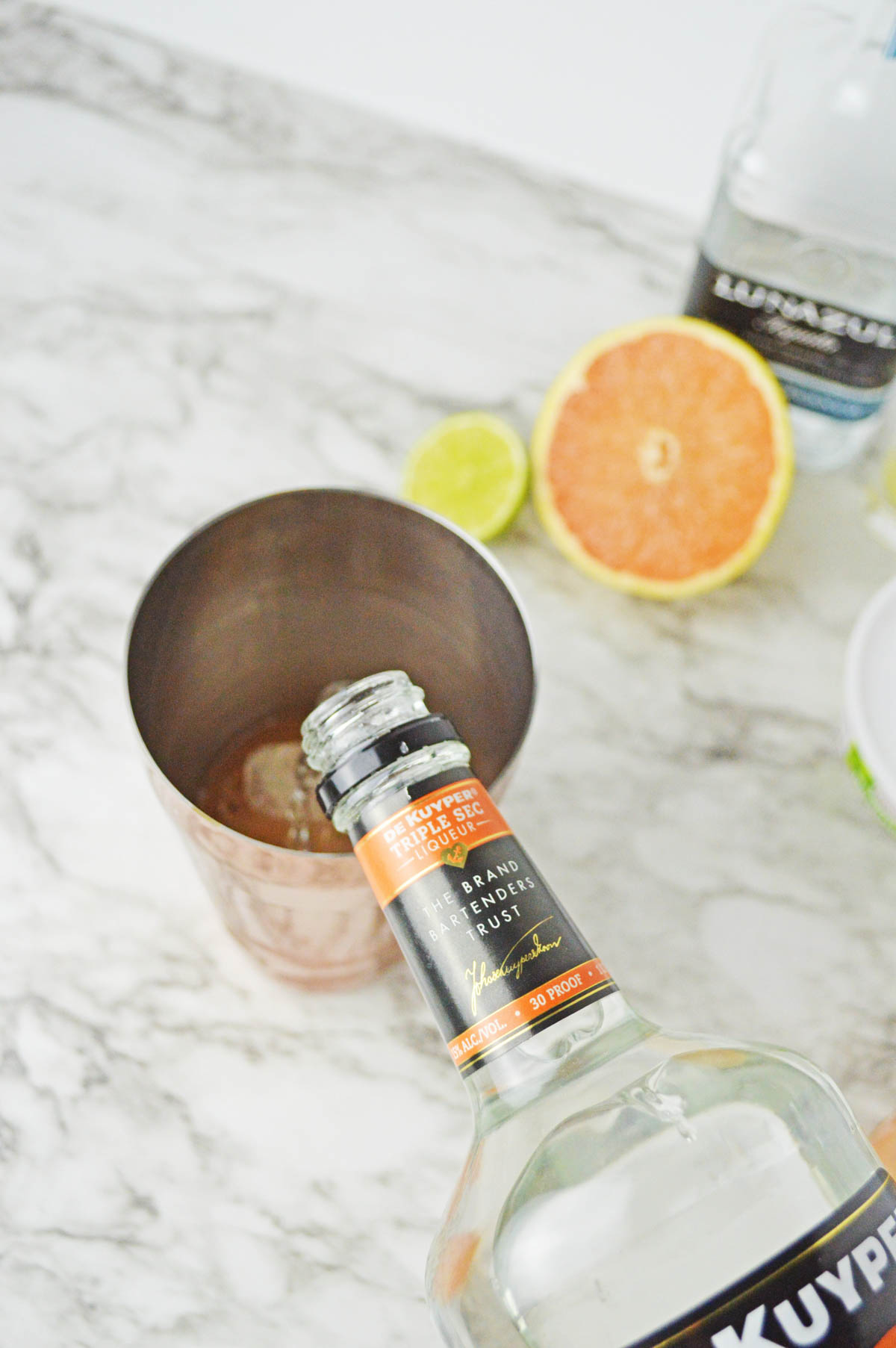 Pouring orange liqueur into a shaker with grapefruit, and lime in background.