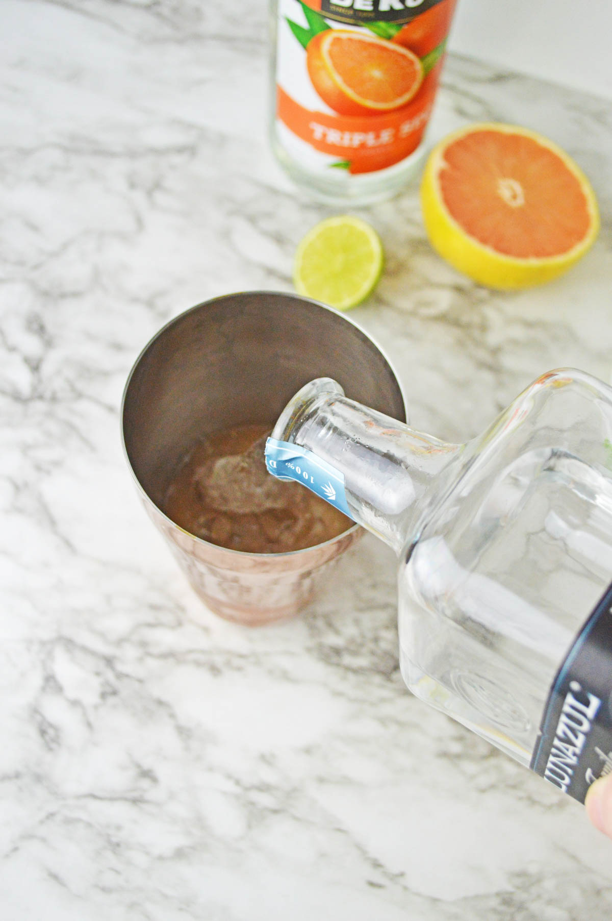 Pouring tequila into a cocktail shaker with orange liqueur, grapefruit, and lime in background.