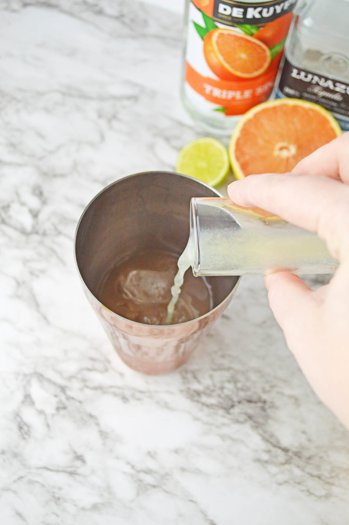 Pouring lime juice into a shaker with orange liqueur, grapefruit, and lime in background.