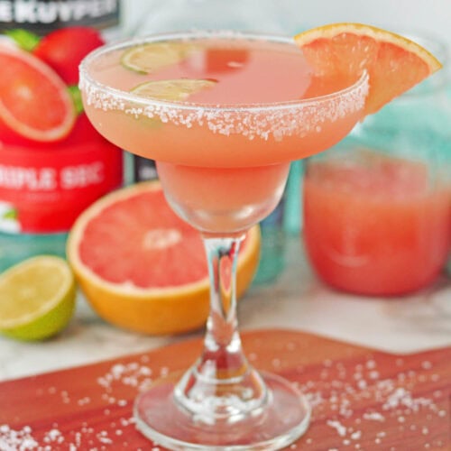 Cocktail set on a wood board made with grapefruit juice in a margarita glass rimmed with salt with lime slices in drink and grapefruit slice on side of rim. Bottle of liqueur in background.