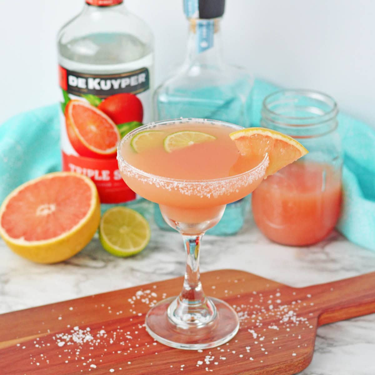 Cocktail set on a wood board made with grapefruit juice in a margarita glass rimmed with salt with lime slices in drink and grapefruit slice on side of rim. Bottle of liqueur in background.