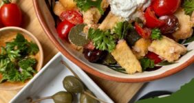 Bowl with salad of tomatoes, capers, shallots, olives, parsley, and dressing topped with feta cheese mixture and caperberry on graphic for Pinterest.
