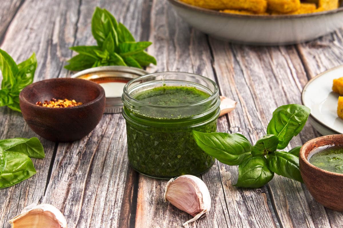 Basil olive oil dipping sauce in a small mason jar with fresh basil, garlic, and wooden bowl of pepper flakes.