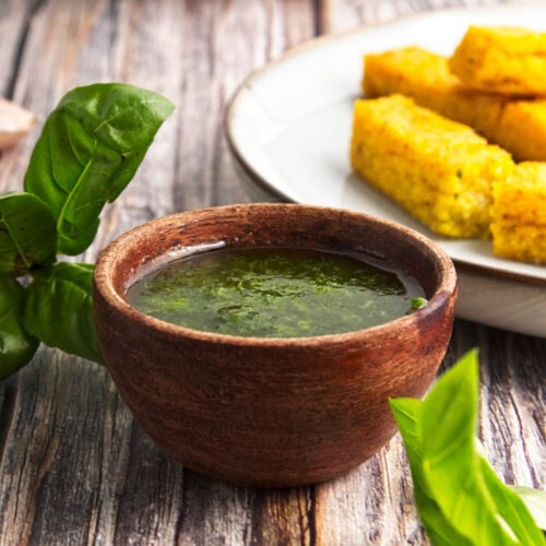 Basil olive oil dipping sauce in a small wooden bowl with fresh basil and bread sticks in the background.