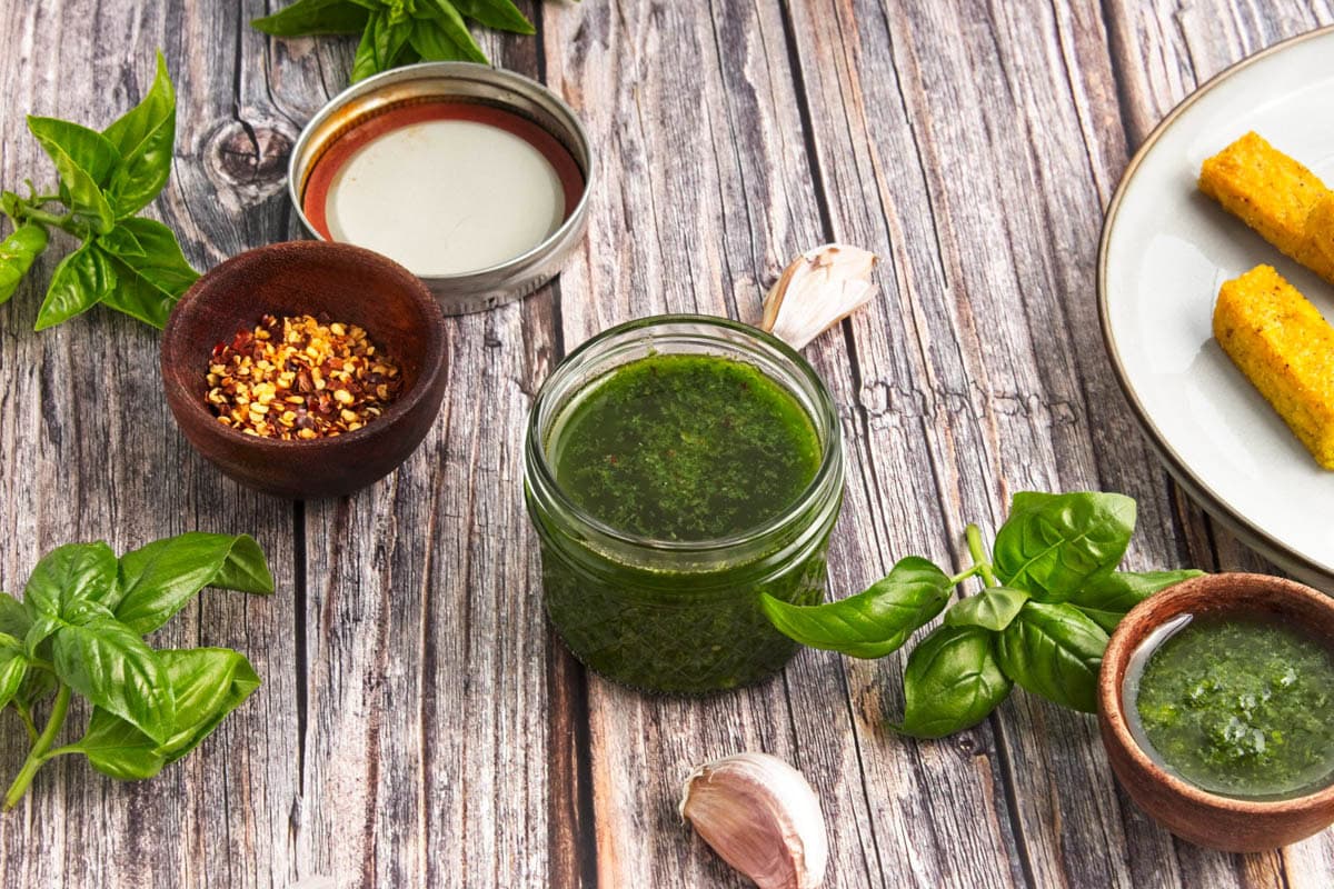 Basil olive oil dipping sauce in a small mason jar with fresh basil, garlic, and wooden bowl of pepper flakes with bread sticks to the side.