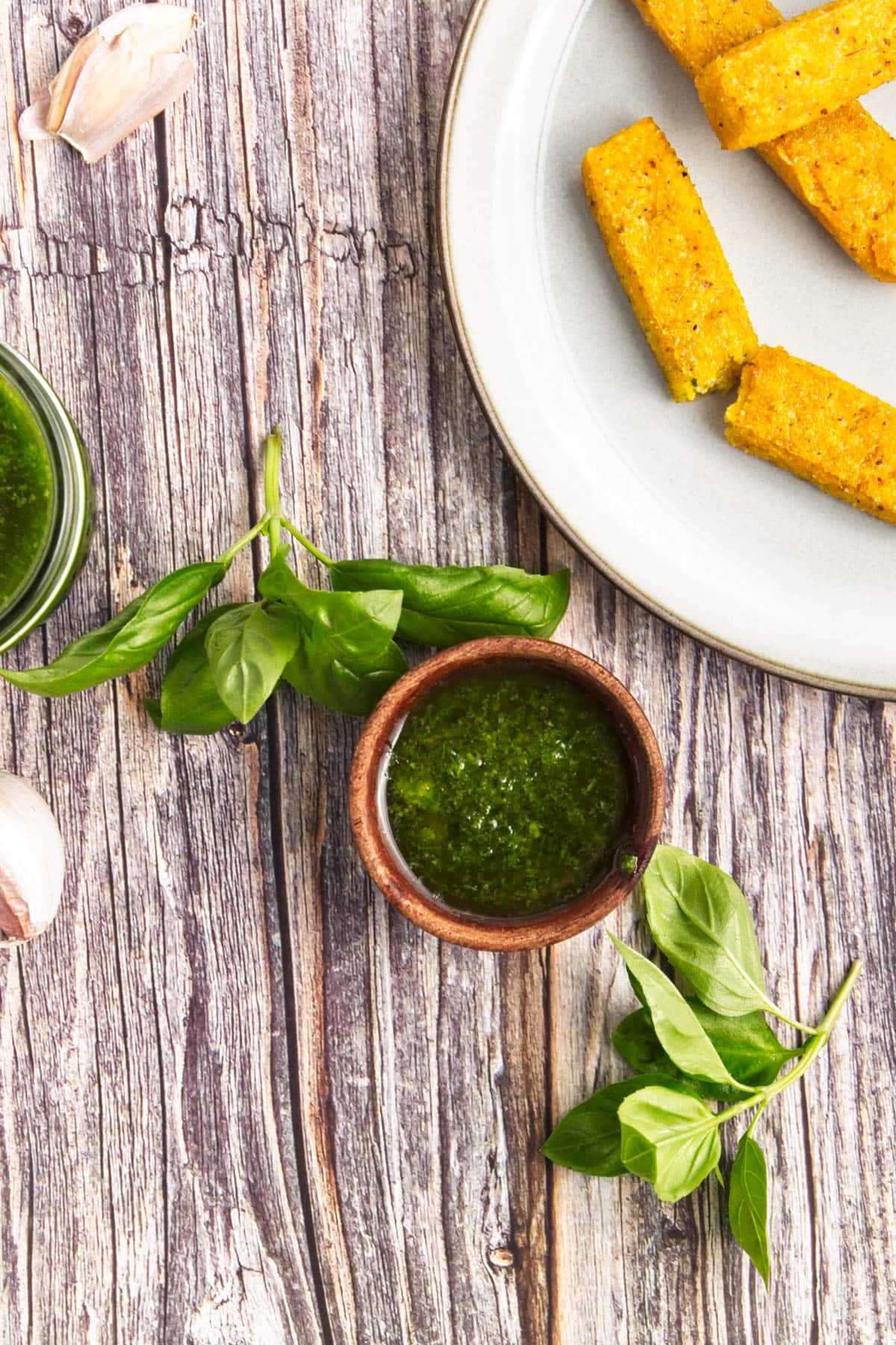 Basil olive oil dipping sauce in a small wooden bowl with fresh basil, garlic, and bread sticks on white plate to the side.