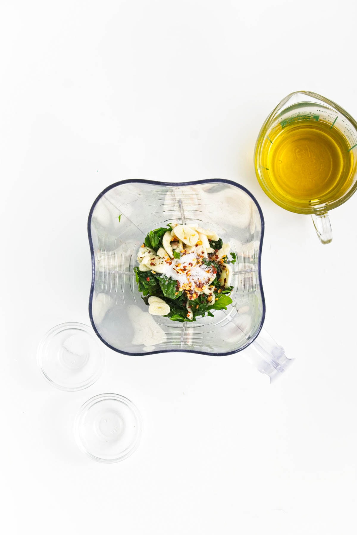 Blender with garlic, basil, salt, and pepper flakes with cup of olive oil to the side.