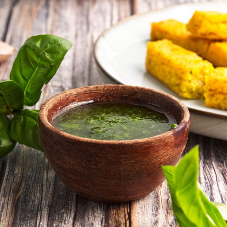 Basil olive oil dipping sauce in a small wooden bowl with fresh basil and bread sticks in the background.