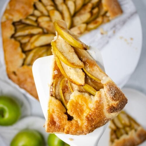 Slice of apple pastry in a crust baked golden brown with rest of pastry below on white platter with green apples in background.