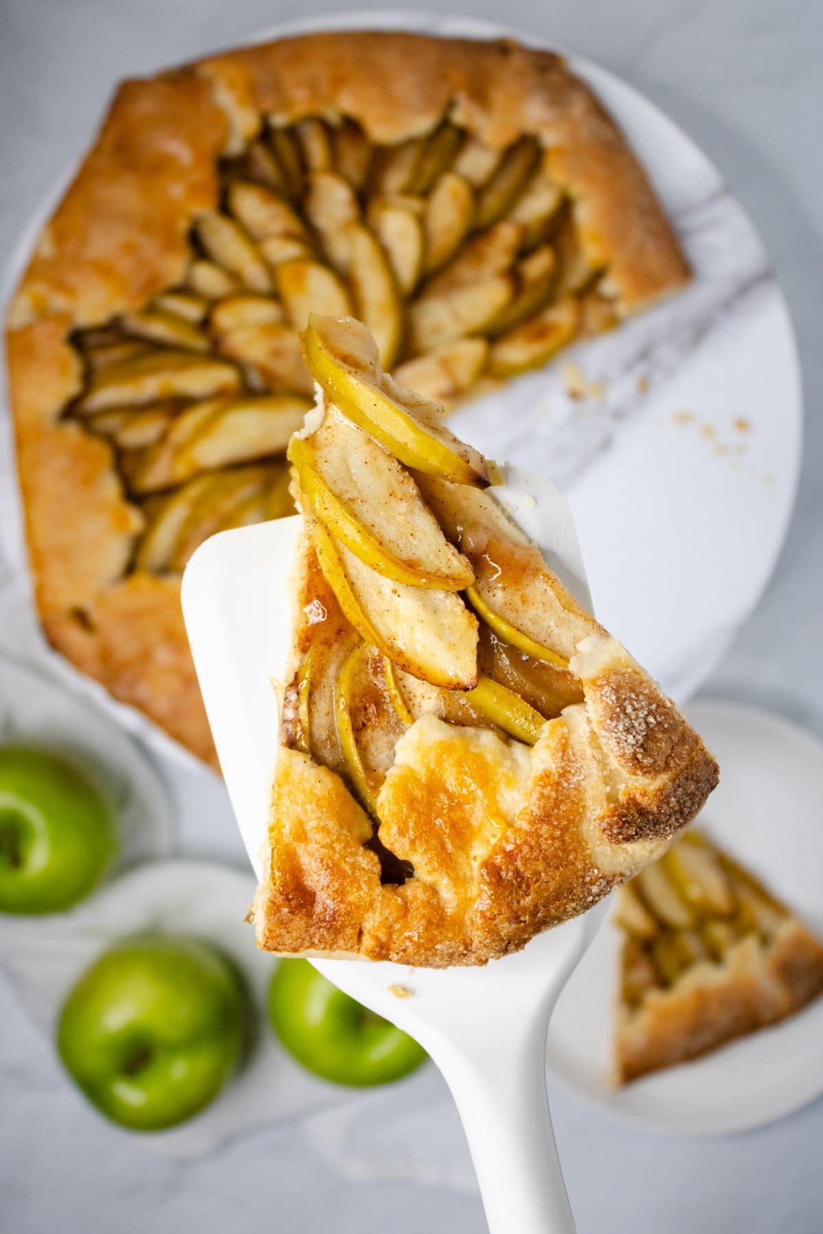 Slice of apple pastry in a crust baked golden brown with rest of pastry below on white platter with green apples in background.