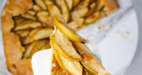 Slice of apple pastry in a crust baked golden brown with rest of pastry below on white platter with green apples in background.