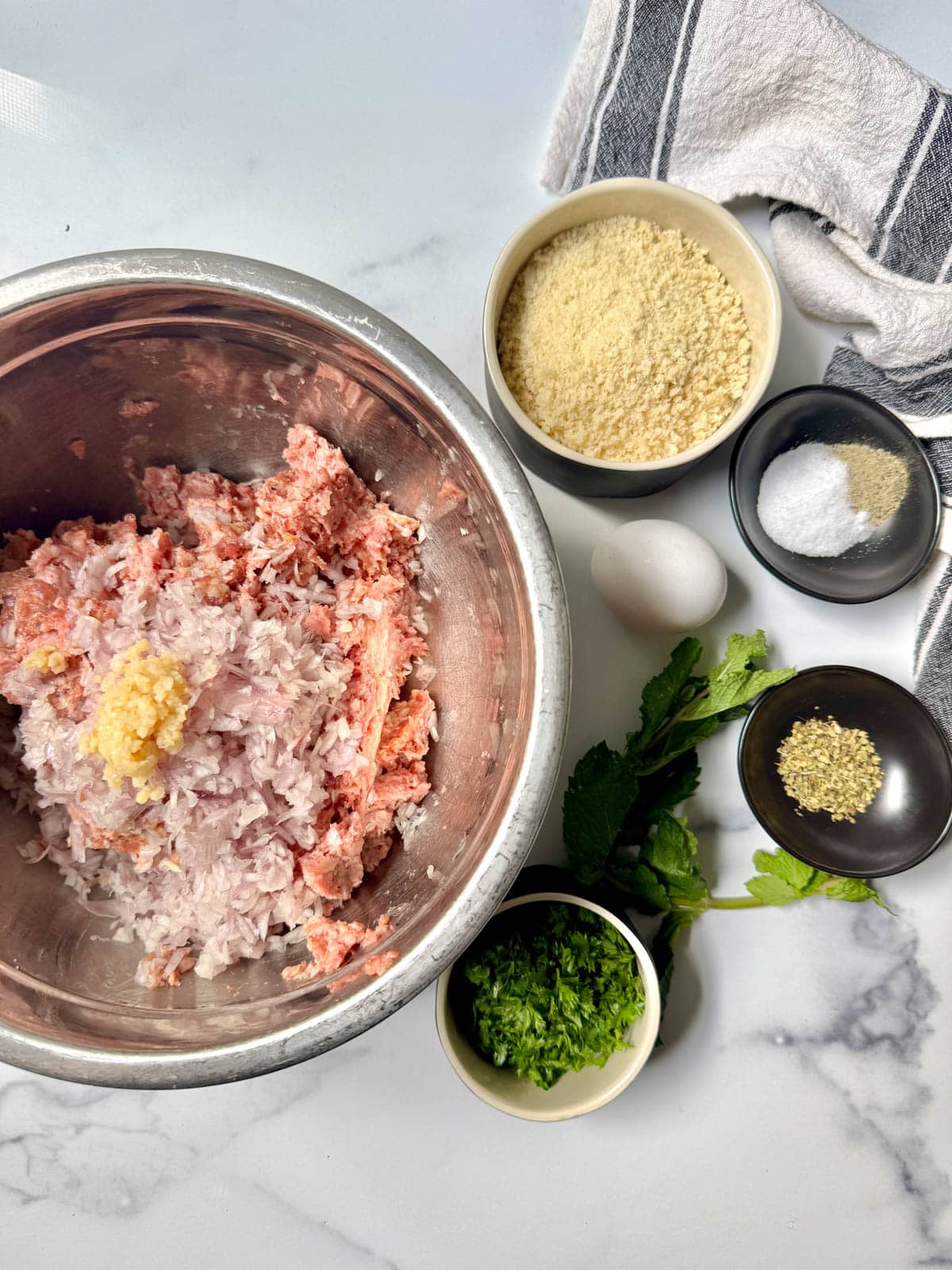 Ground meat, shallot, and garlic in bowl with small bowls of seasonings, bread crumbs, and an egg.