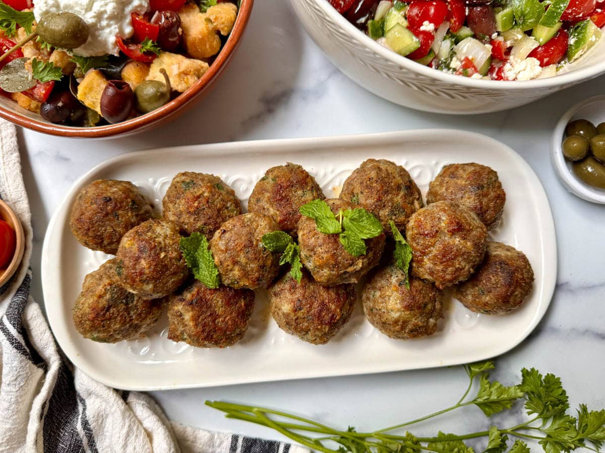 Greek meatballs on a white platter with parsley, tomatoes, and a salad to the side.