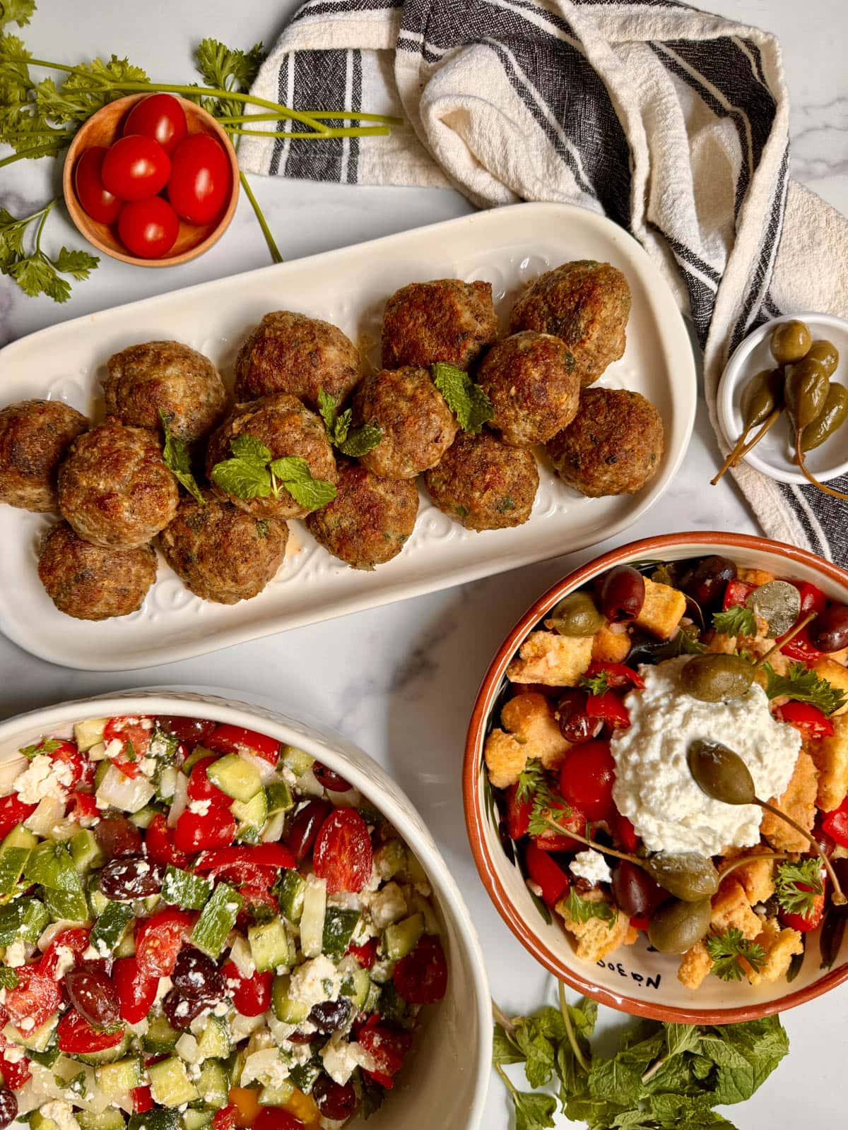 Greek meatballs on a white platter with parsley, tomatoes, and a salad to the side. with small bowl of tomatoes and parsley above, small bowl of caperberries, and two salads below.