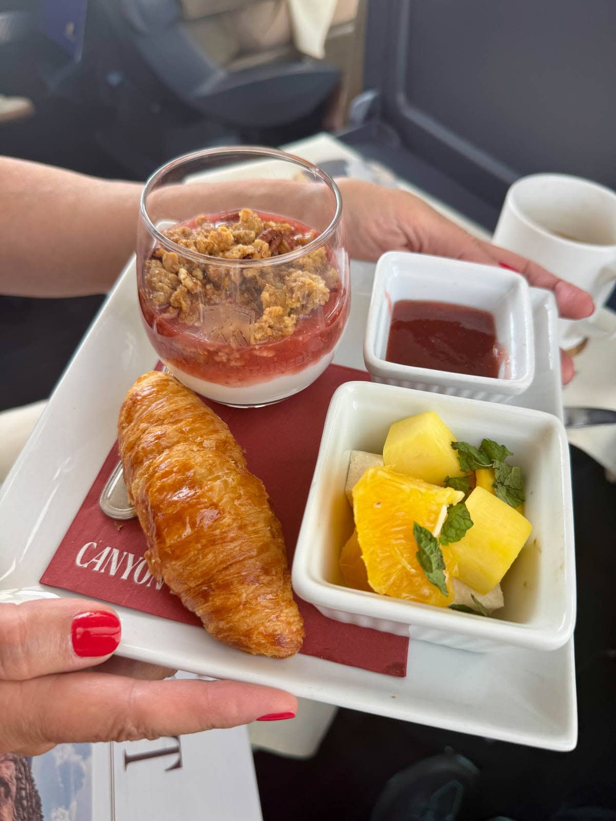 Yogurt parfait in a glass, a croissant, and a bowl of fruit with a coffee cup to the side.
