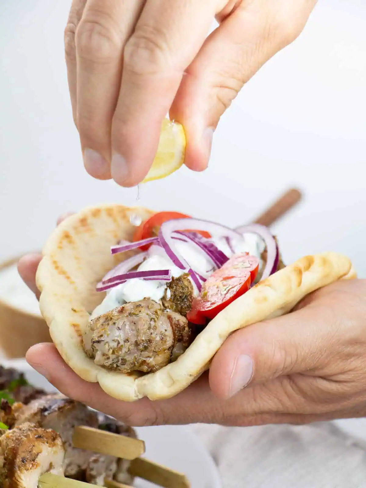 Chicken skewer in pita bread with sauce, onion, and tomato with hand squeezing lemon above.