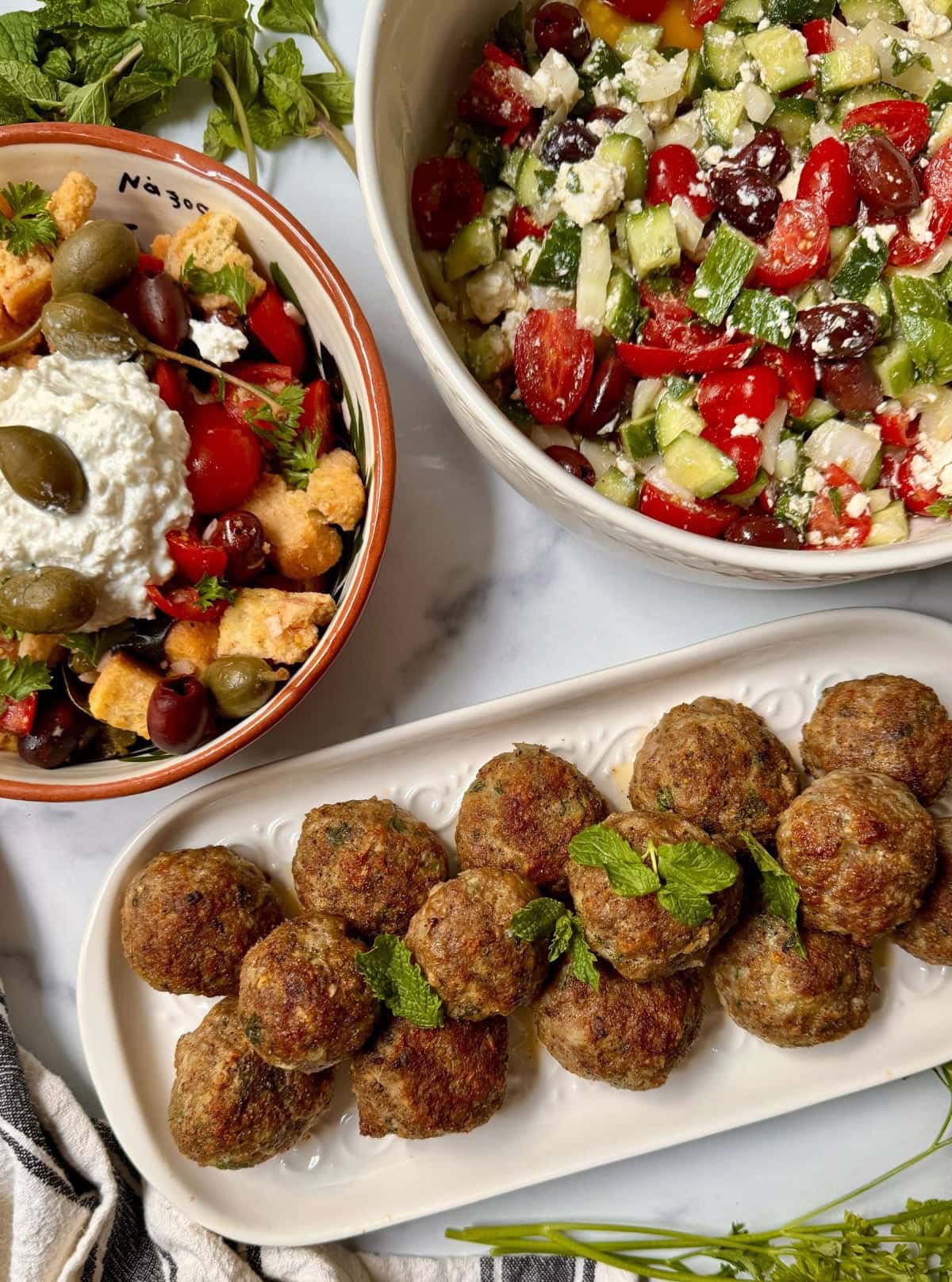 Two Greek salads and a platter of Greek meatballs on a white counter.