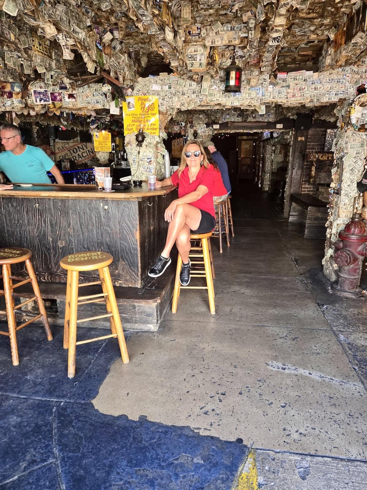 Woman sitting on a wooden barstool at a bar with dollar bills all over the walls.