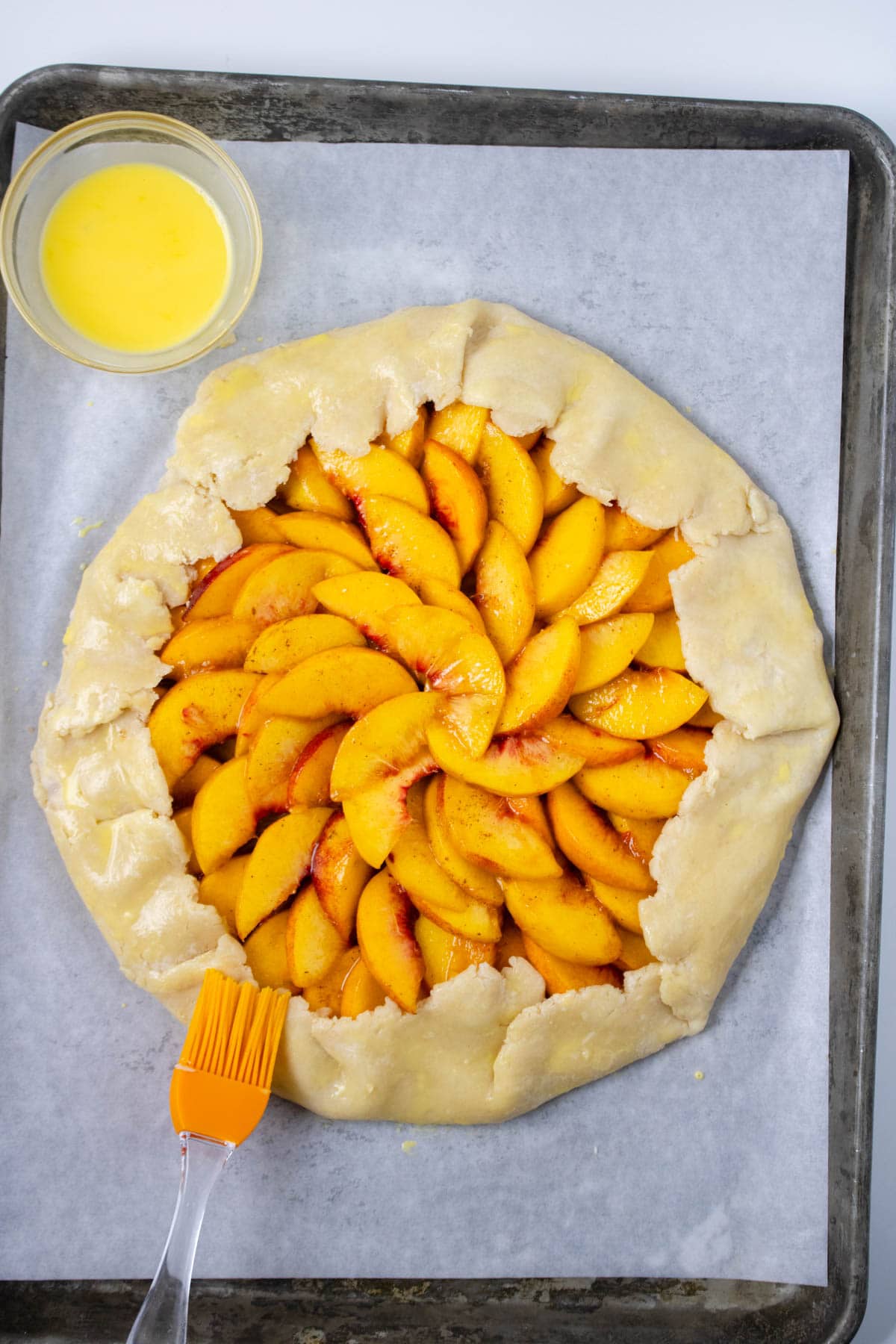 Pie crust rolled out and set on parchment paper on baking sheet topped with sliced peaches with part of crust folded over fruit and adding an egg wash.