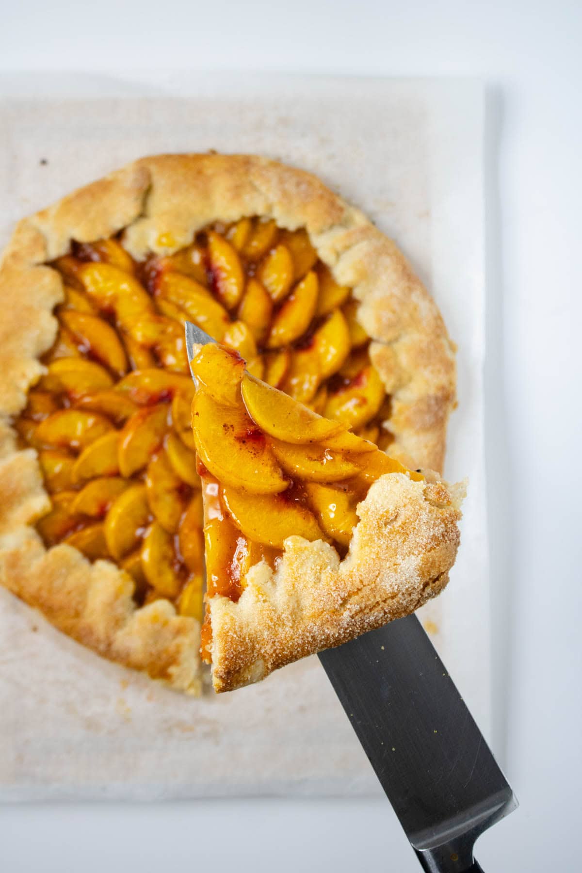 Baked peach galette on parchment paper with one slice pulled out and held above on a knife.